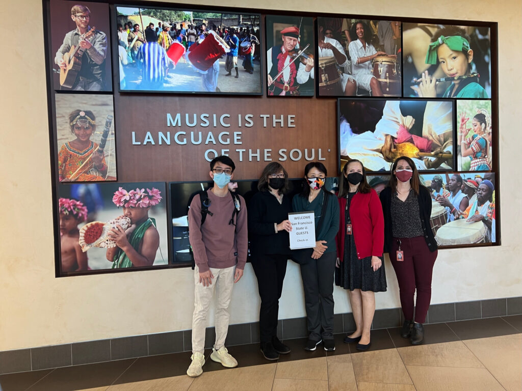 Leigh Jin, Weimin Zhang, and Anh Tran with Music History Museum staff in front of the a mural with a welcome sign. 