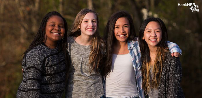 Four young women of diverse ethnic backgrounds with their arms around each other smiling. 